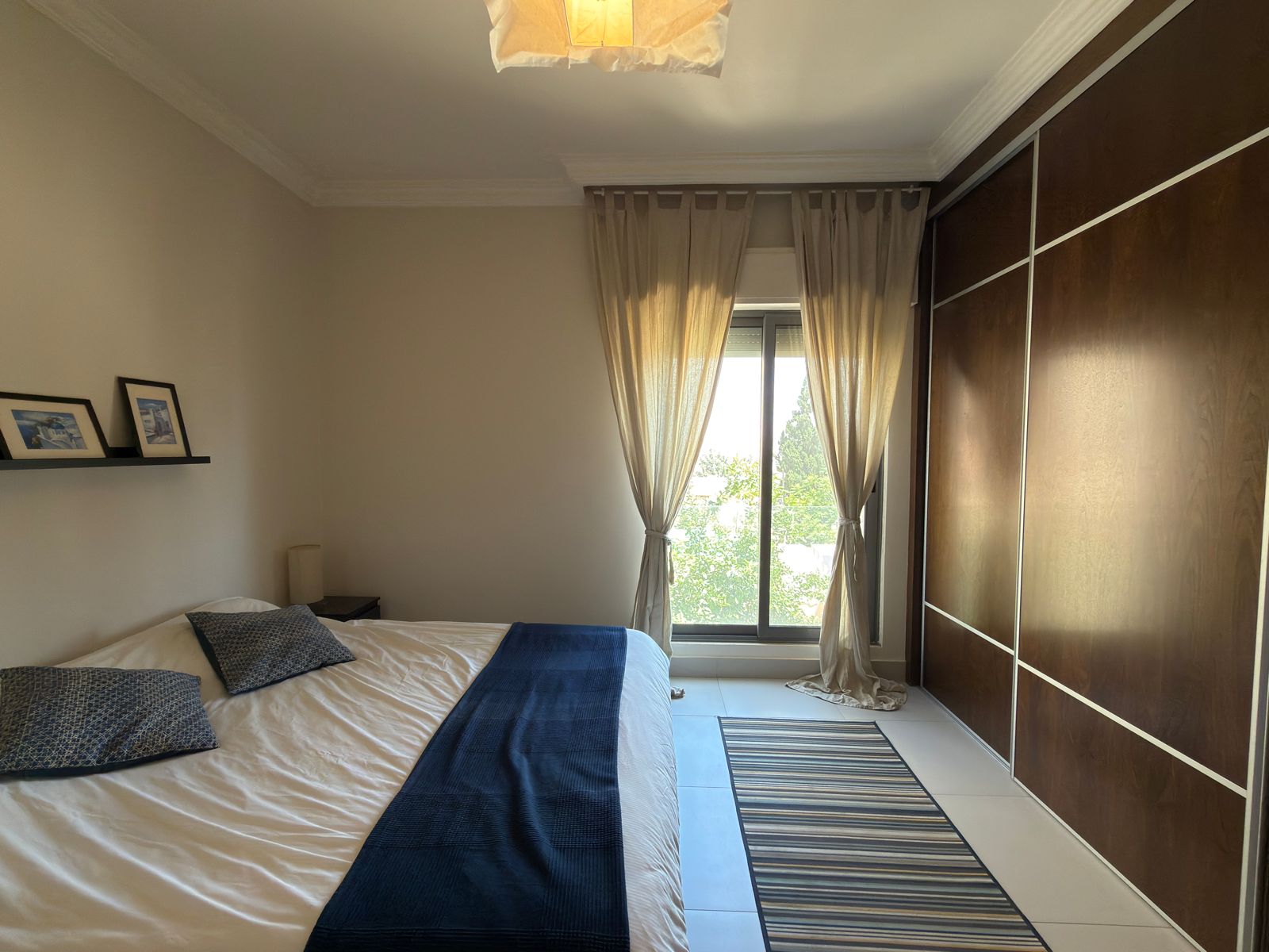 45979b27-78fb-40b8-b637-04ccab4167ea Bedroom with a made bed, navy runner, and patterned pillows by a window with sheer beige curtains; a large dark wood wardrobe lines the opposite wall.