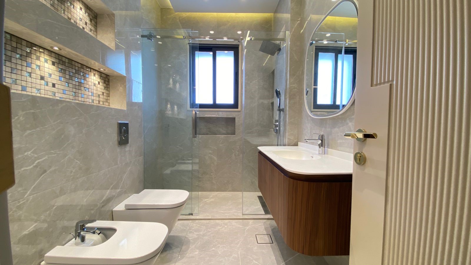 45e50557-b3d8-424b-a924-0d2549662a2d Modern bathroom with glass shower enclosure, wall-to-wall marble tiles, and a wooden vanity with white countertop and oval mirror far wall window above. Includes toilet and bidet.