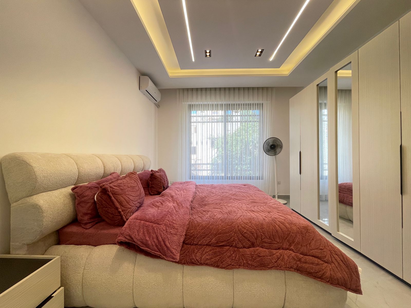 46151917-db62-41df-98f7-85eb1eaed1ca Bright modern bedroom with a large beige upholstered bed and maroon quilted bedding. Wardrobe with mirrored doors along the right wall; ceiling recessed lighting; window with sheer curtains in the back; standing fan near the window.