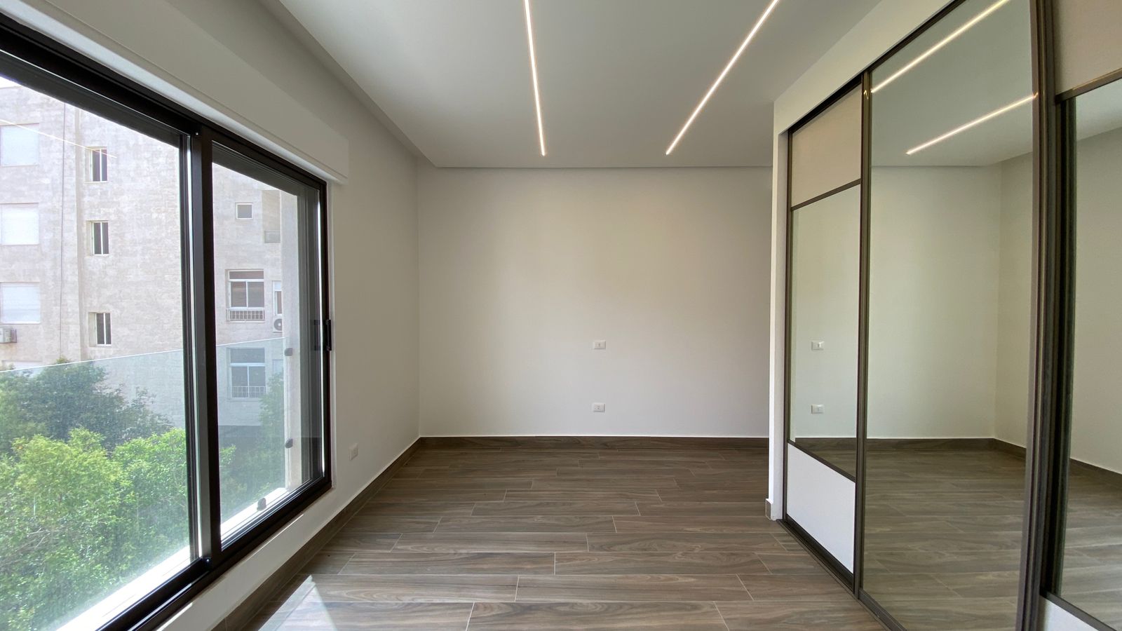 4624c426-97dc-4865-be24-ab92c4c09cf2 Empty modern room with large floor-to-ceiling windows overlooking a neighboring building and greenery, wood-look flooring, and a mirrored wardrobe on the right.