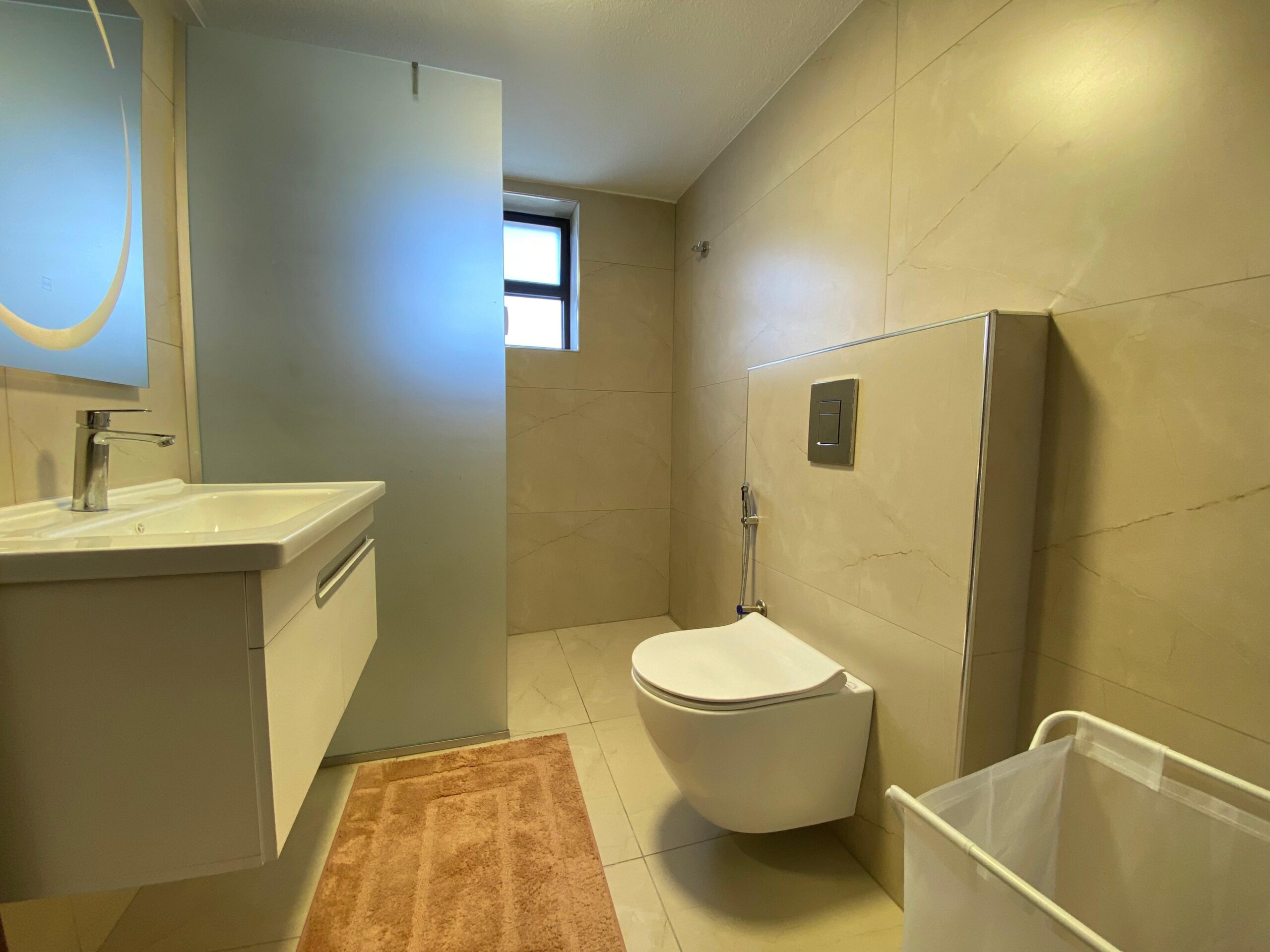 46264c79-aa23-4584-bcc5-602eb75704ce Modern bathroom with white vanity and chrome faucet on the left, beige marble walls, and a wall-hung toilet on the right side.