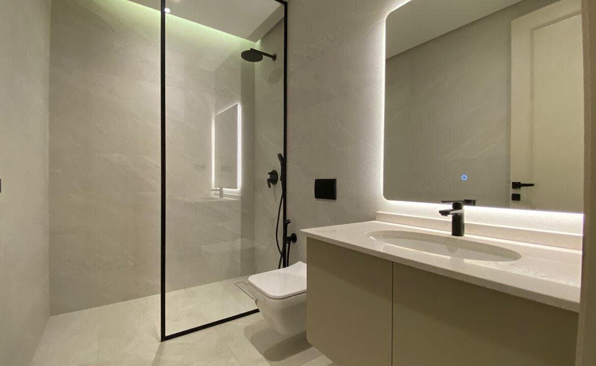 Modern bathroom with a glass-enclosed shower, white vanity, and backlit mirror in a minimalist gray space.