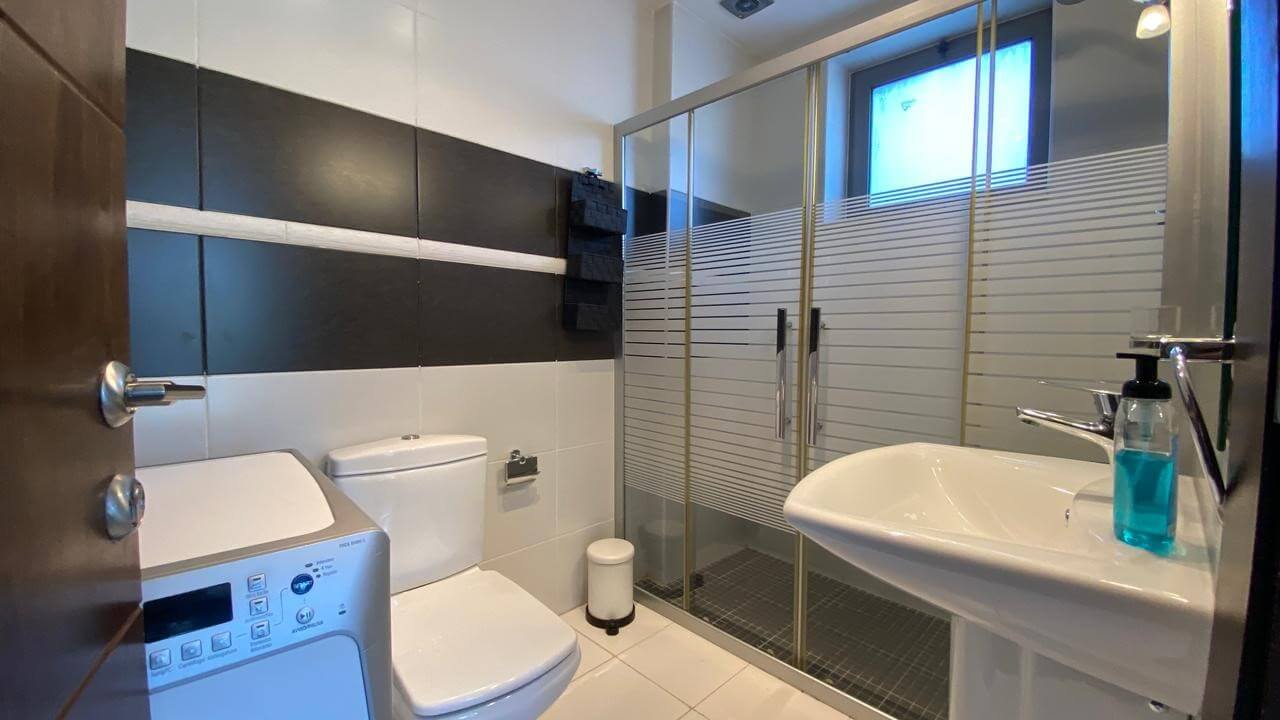 473ac7ce-9ae5-4401-9f1a-0315627bcd2a Modern bathroom with washing machine, toilet, pedestal sink, and a glass shower enclosure with frosted stripes; blue liquid soap on sink.