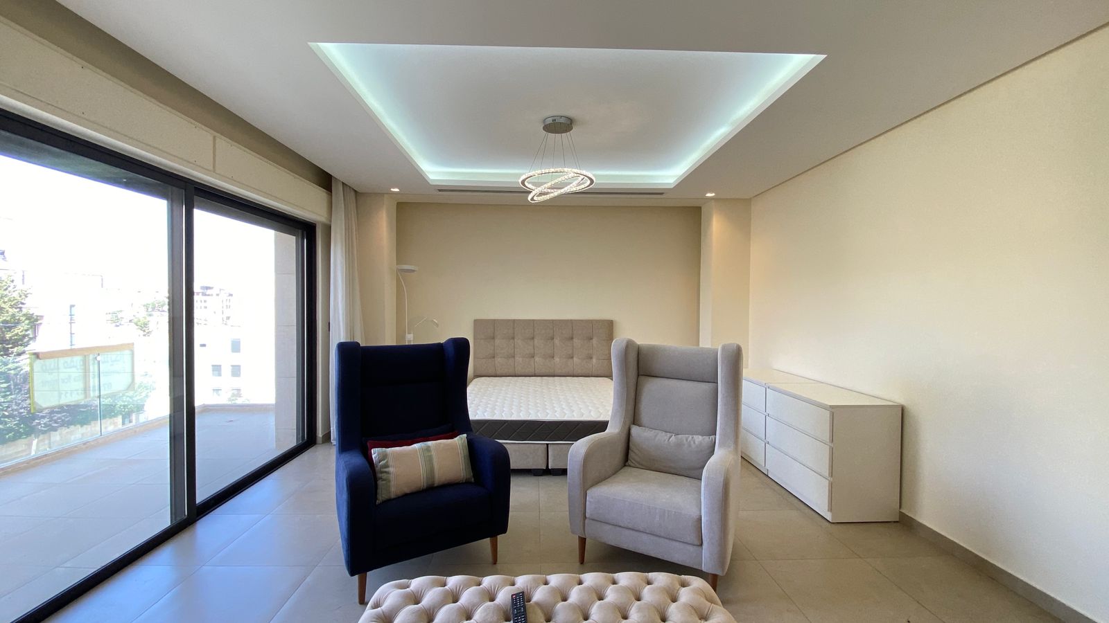 47c097c1-3f14-4afe-bf37-0f01dc228cd5 Bright modern living area with two armchairs, a tufted ottoman, and a bed in the background; sliding glass doors open to a balcony on the left.