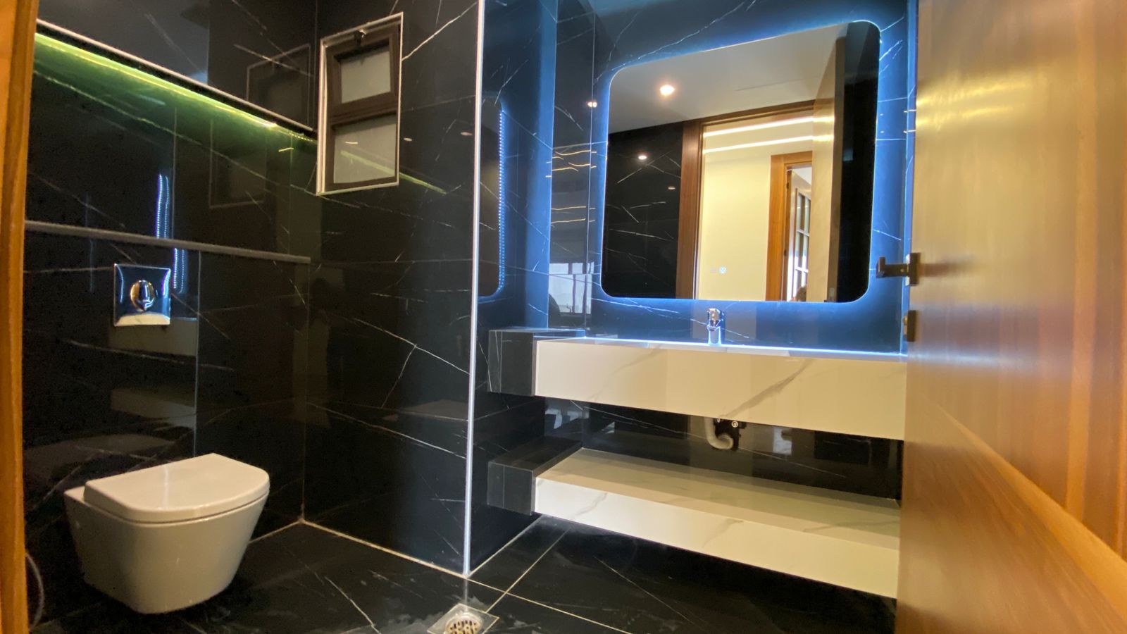 485738ae-2ba3-4e8a-82ab-3370e79e799f Modern bathroom with black marble walls, wall-hung toilet, and a blue-lit rectangular mirror above a white sink counter.