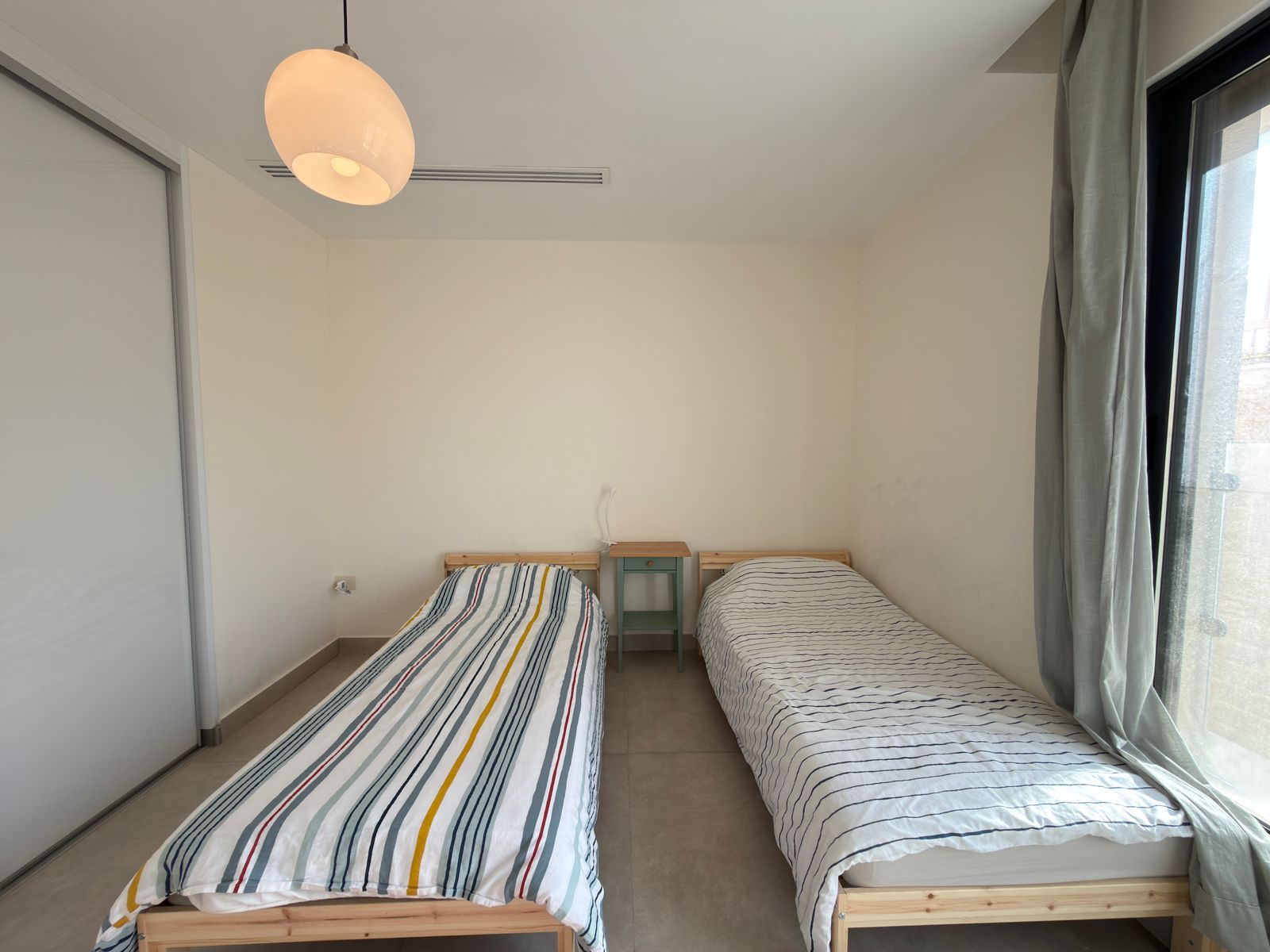 4917d97e-9e93-4cc2-8357-ac06598370db Small bedroom with two single beds and a shared nightstand between them, white walls, and a large window with gray curtains.