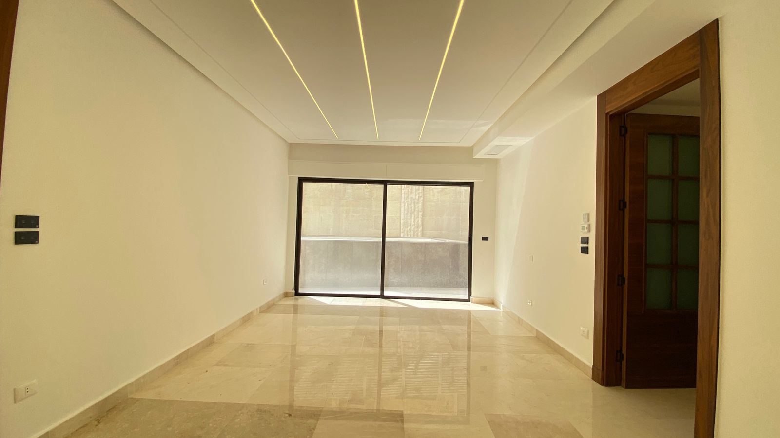 4995e9f7-899b-4f84-9c75-67aac0858f15 Empty living room with polished marble floor, recessed ceiling lights, and a sliding glass door to a balcony.