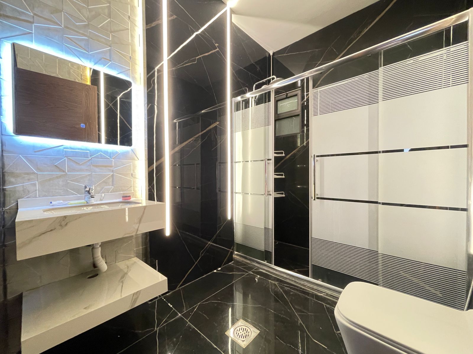 49bdb1b1-b2ac-44ff-ab1c-1a5e5279798d Luxurious bathroom with black marble walls, a backlit mirror above a white wall-mounted sink, and a frosted-glass shower enclosure.