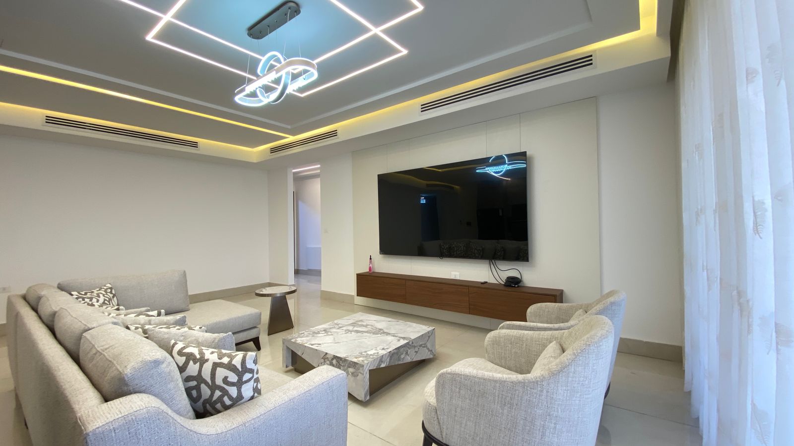 49e0d4d2-a564-4713-8540-007af1008bb4 Modern living room with a wall-mounted flat-screen TV above a wooden console, gray sectional sofa, and two matching armchairs around a marble coffee table.