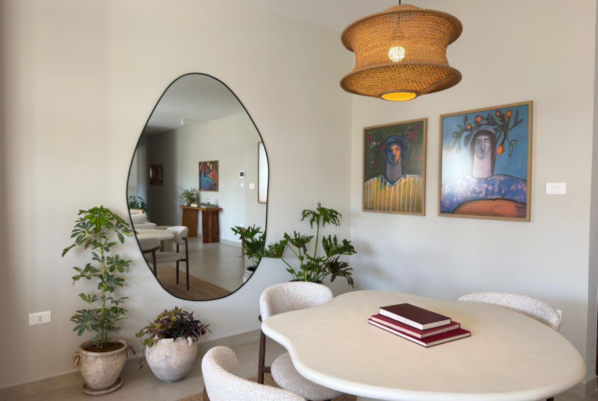 A round white dining table with beige upholstered chairs, two red books stacked on top, in a bright room with potted plants and a large oval wall mirror reflecting a sofa and artwork in the background. A woven pendant light hangs from the ceiling and two colorful portraits are mounted on the wall.