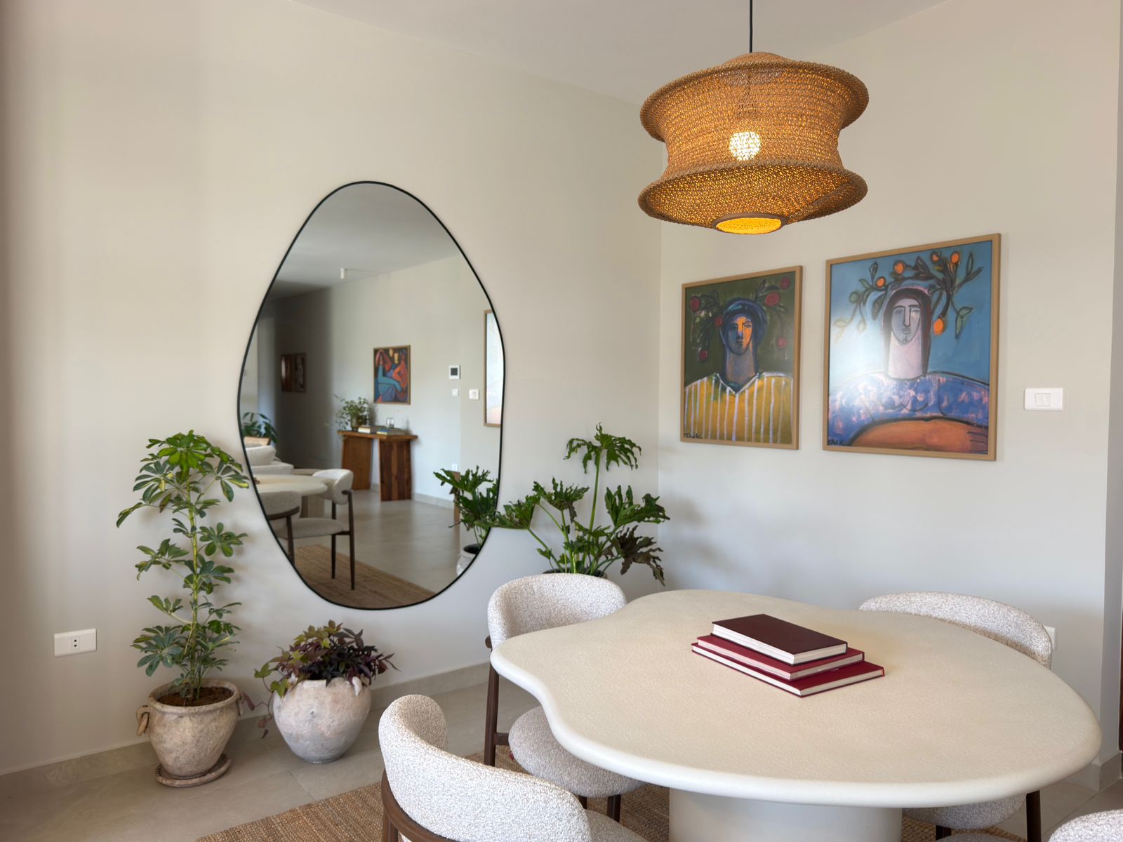 4a1fb30c-1e41-46c3-8458-3cad18fe9a7a A round white dining table with beige upholstered chairs, two red books stacked on top, in a bright room with potted plants and a large oval wall mirror reflecting a sofa and artwork in the background. A woven pendant light hangs from the ceiling and two colorful portraits are mounted on the wall.