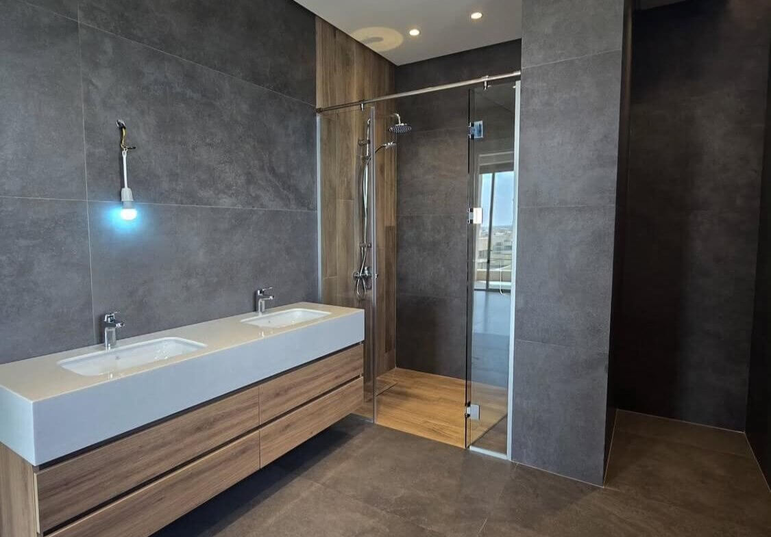 4a30a42b-a5c2-4f1b-8e5c-fb5d8c302b9c Modern bathroom with a white double-sink vanity, wood drawers, gray tile walls, and a glass shower. (informative)