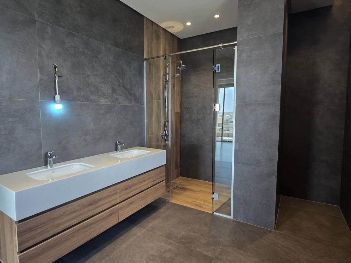 4a30a42b-a5c2-4f1b-8e5c-fb5d8c302b9c Modern bathroom with a white double-sink vanity, wood drawers, gray tile walls, and a glass shower. (informative)