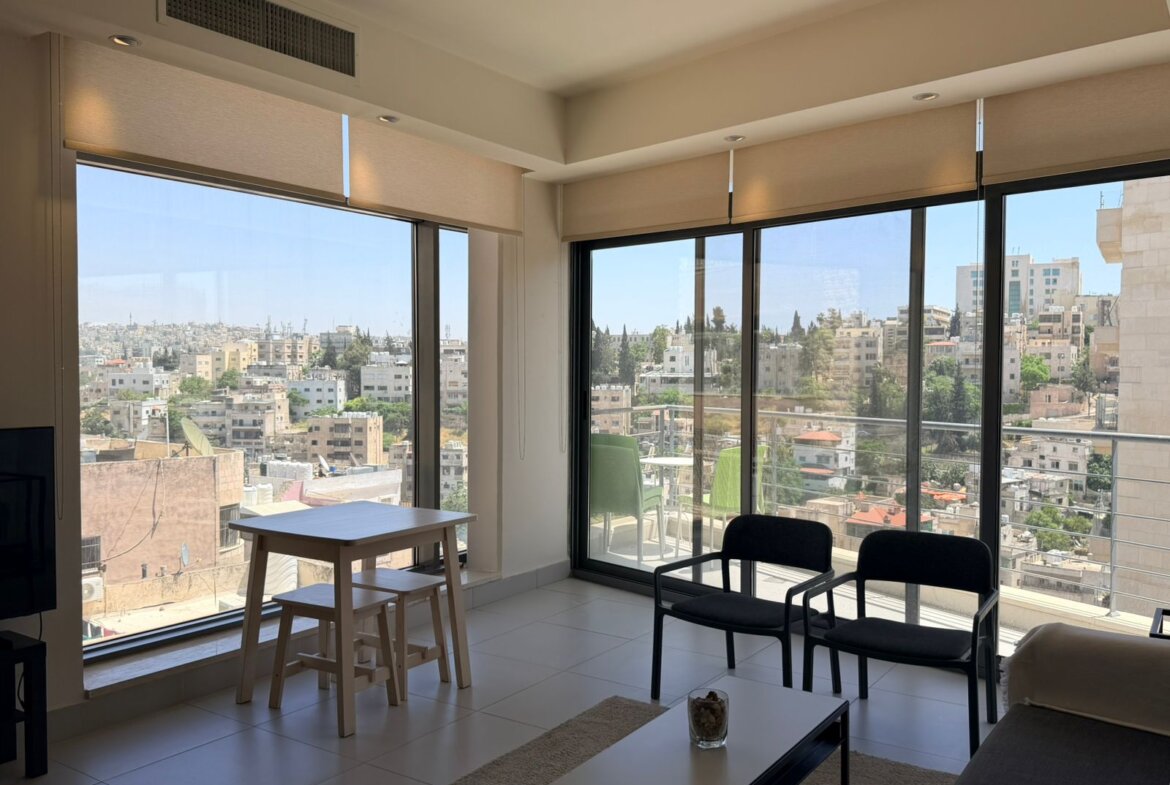 Bright living room with floor-to-ceiling windows showing a cityscape and balcony; light beige roller blinds partially down and contemporary seating.