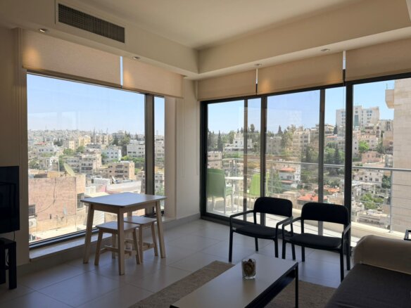 Bright living room with floor-to-ceiling windows showing a cityscape and balcony; light beige roller blinds partially down and contemporary seating.