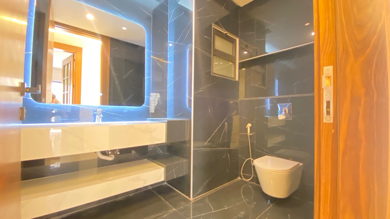 4a8e32d1-8780-401c-9e56-ea6ebec173ac Modern bathroom with black marble walls, a backlit rectangular mirror above a white sink and countertop, and a wall-mounted toilet.