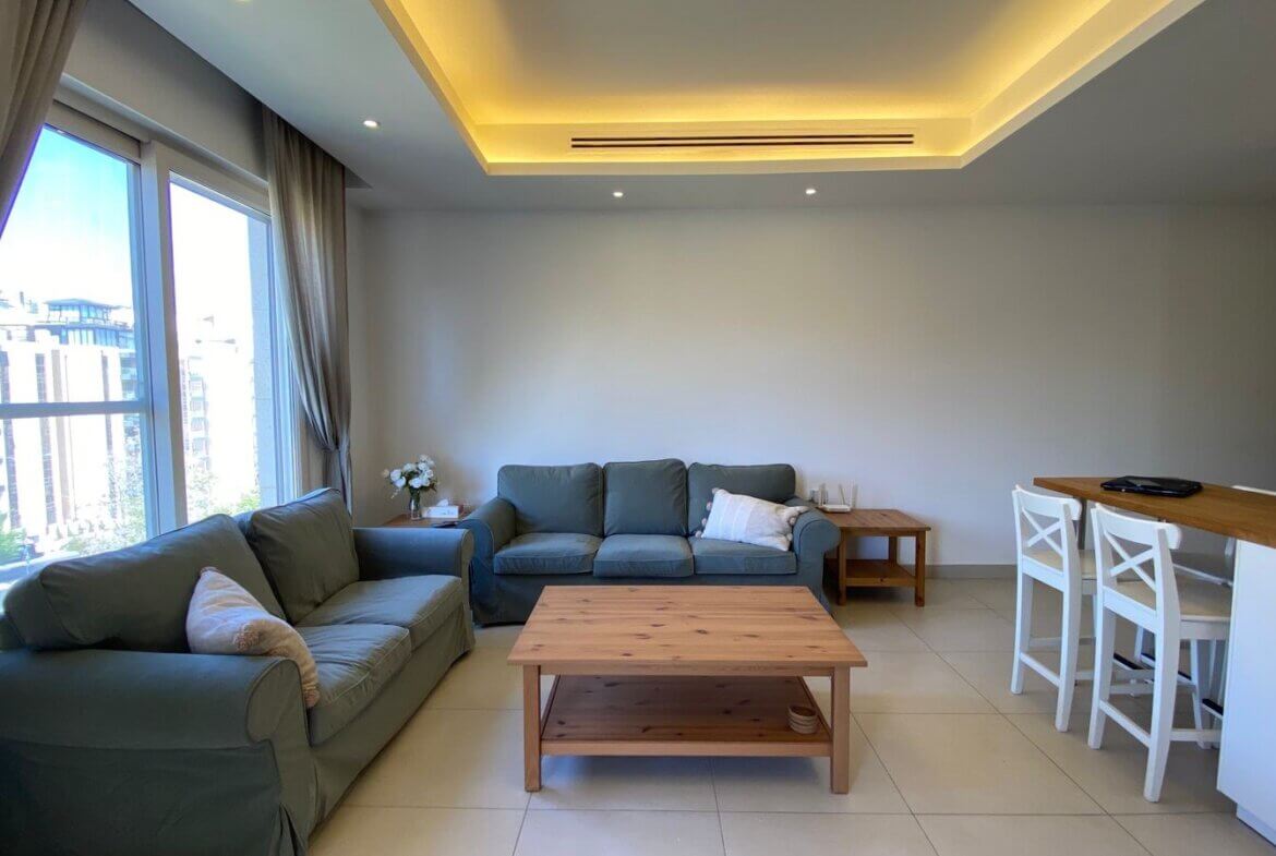 Bright living room with two teal-green sofas, a wooden coffee table, and a side table against a light gray wall; large windows with beige curtains reveal an urban view.