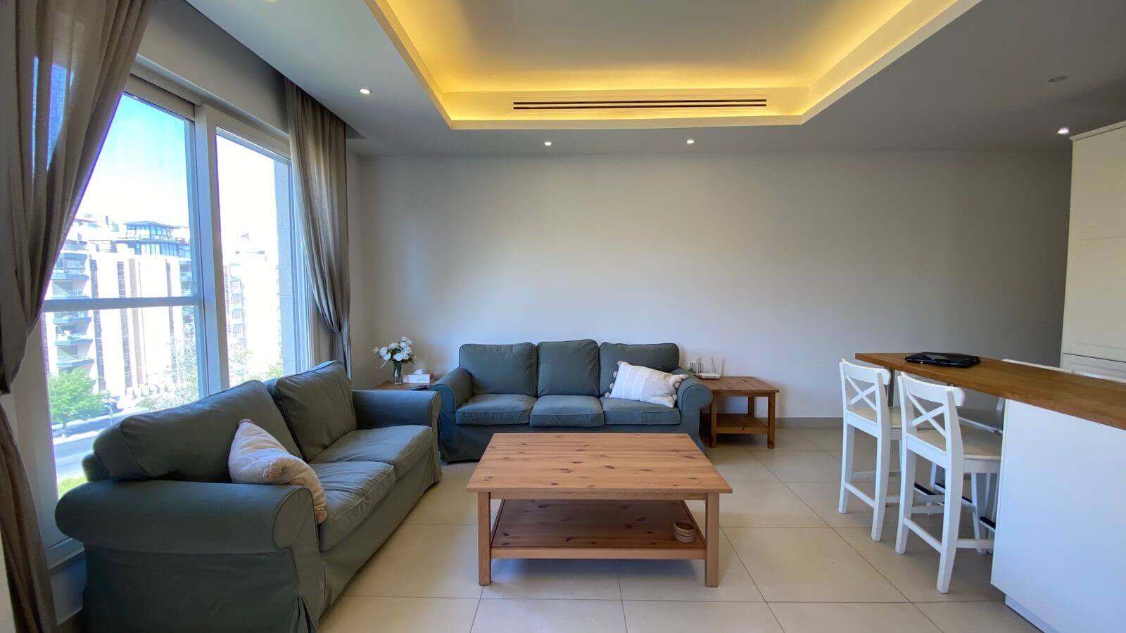 4b34a3ee-abf9-4714-b7c5-b81b58f92f80 Bright living room with two teal-green sofas, a wooden coffee table, and a side table against a light gray wall; large windows with beige curtains reveal an urban view.