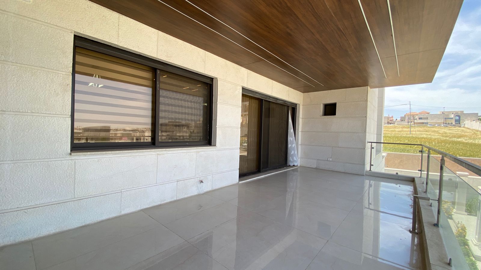 4c50d005-fbad-40bd-ae1a-d9e16ffe4bfd Empty modern balcony with white stone walls, large sliding glass doors, and a glass railing overlooking a suburban area.