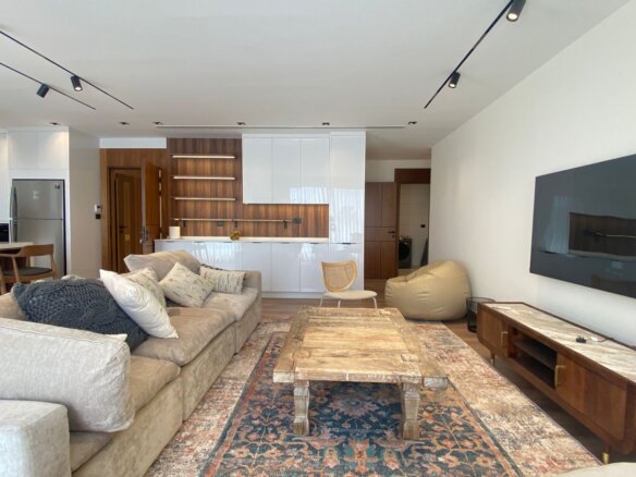 Open-concept living room with beige sectional, rustic wooden coffee table on a patterned rug, and a wall-mounted TV; kitchen area with white cabinets and bar stools in the background.