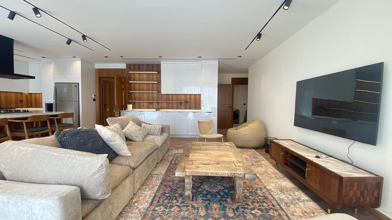 4c999fec-65c8-48cc-9d7f-53bad5414f2d Open-concept living room with beige sectional, rustic wooden coffee table on a patterned rug, and a wall-mounted TV; kitchen area with white cabinets and bar stools in the background.