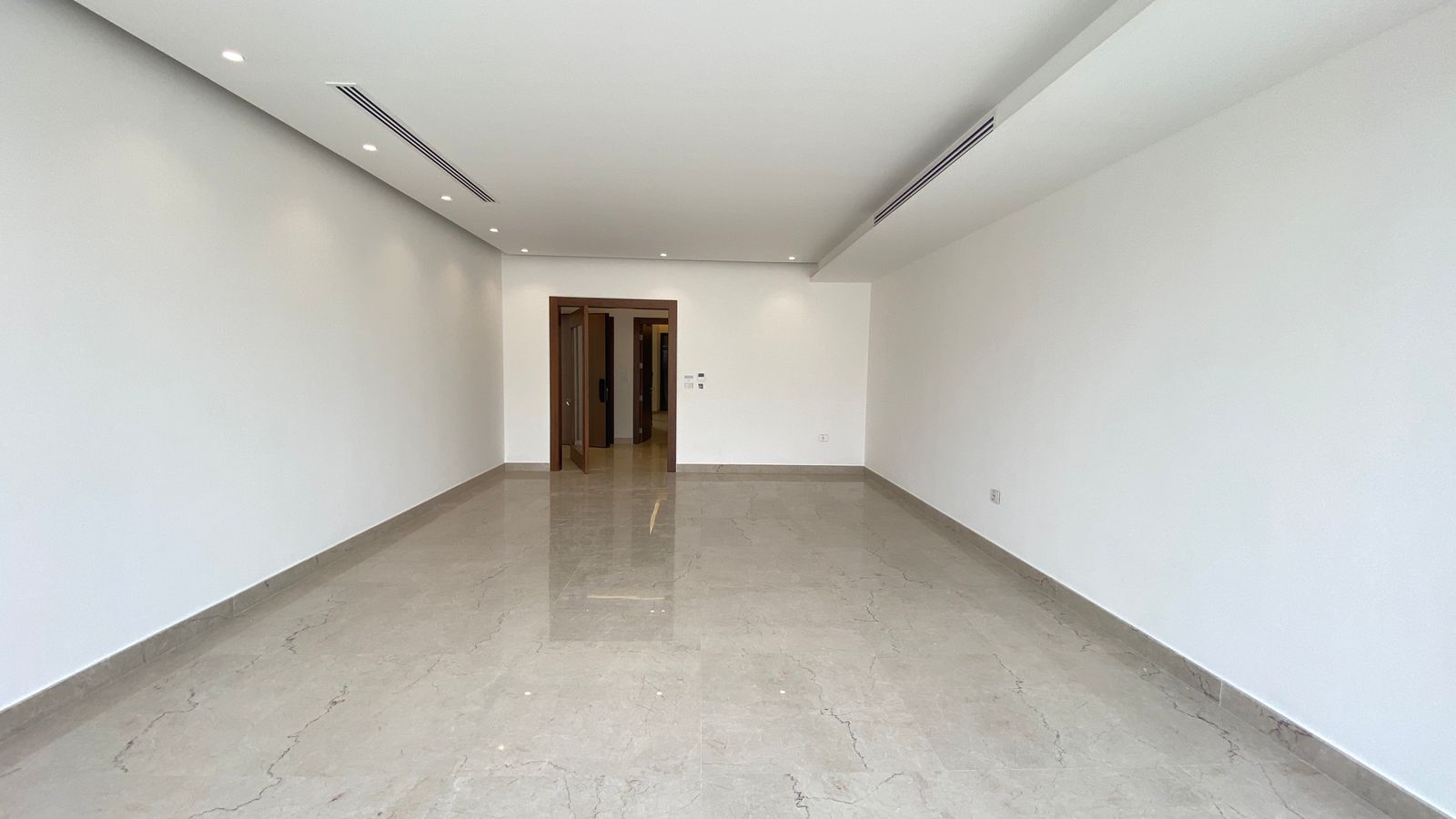 4cc40259-7cec-4338-aef9-adde4a490674 Bright empty commercial space with white walls, glossy beige marble floor, and recessed ceiling lights along the left side; a wooden doorway at the far end opens into another area.
