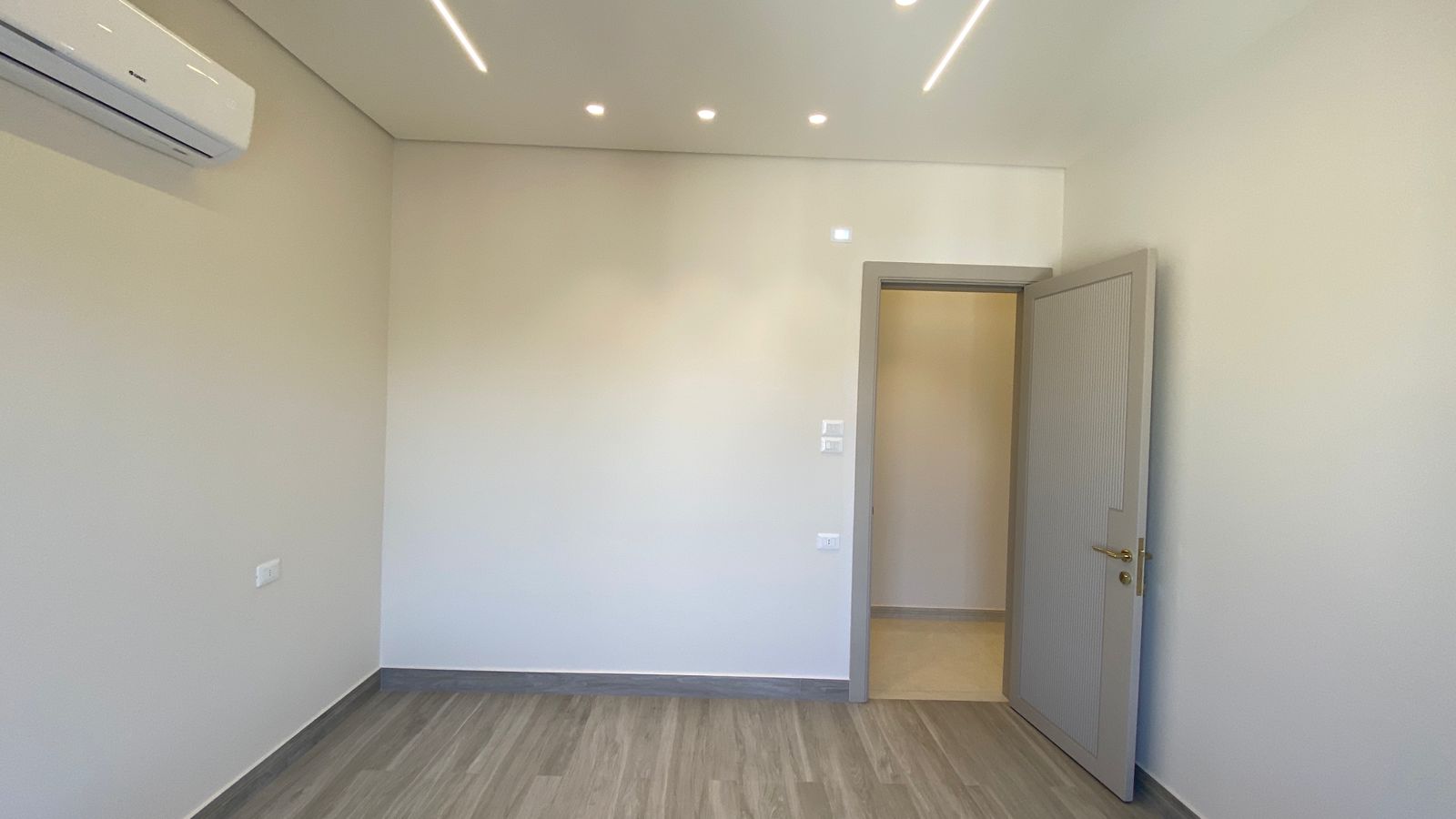 4ce570b0-7439-47b5-97f9-5b5780f48815 Empty modern room with light gray walls, wood-look flooring, ceiling LEDs, and an open door to a hallway.