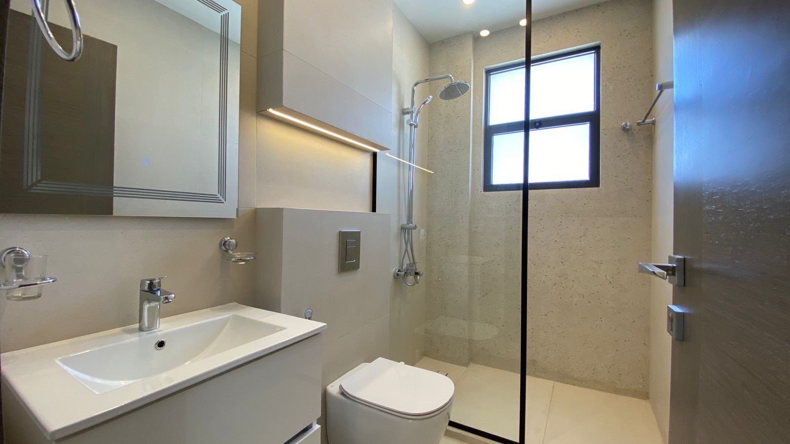 4d2d4040-3ea5-446e-96a9-9967a6cae679 Modern bathroom with white sink, wall mirror, wall-mounted toilet, and a glass-enclosed shower near a frosted window.