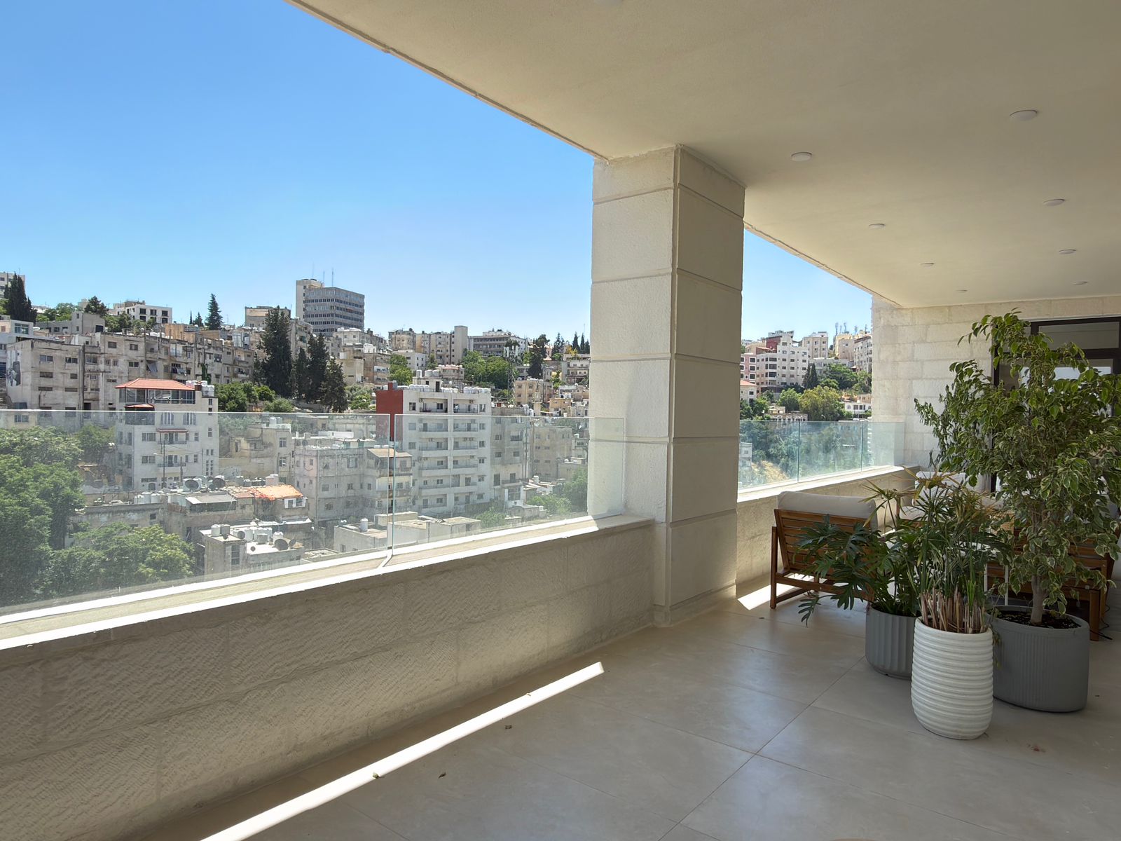 4da3ece7-5eac-4ade-aa97-b8cc1aee7eea Balcony with glass railing and potted plants overlooking a cityscape under a blue sky.