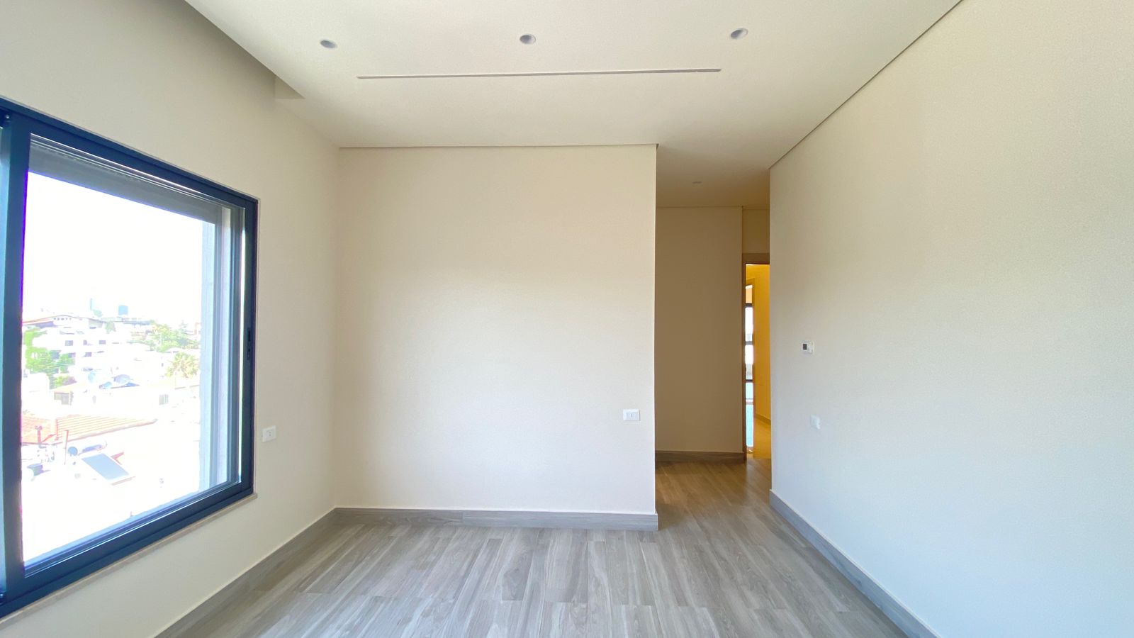 4e4de0ce-8f84-4368-8471-d2c020fca6d2 Empty bright room with light gray wood flooring, white walls, and a large window on the left showing an outdoor view; hallway ahead to a warmly lit space