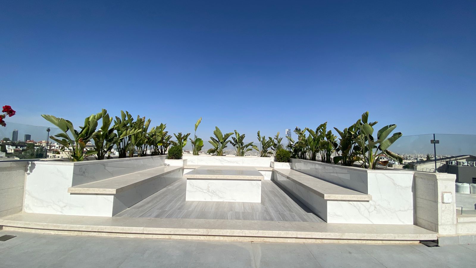 4e8263fe-8a71-4655-8c6c-7504c6b4590a Rooftop seating area with white marble benches and planters, lined with banana plants under a clear blue sky.