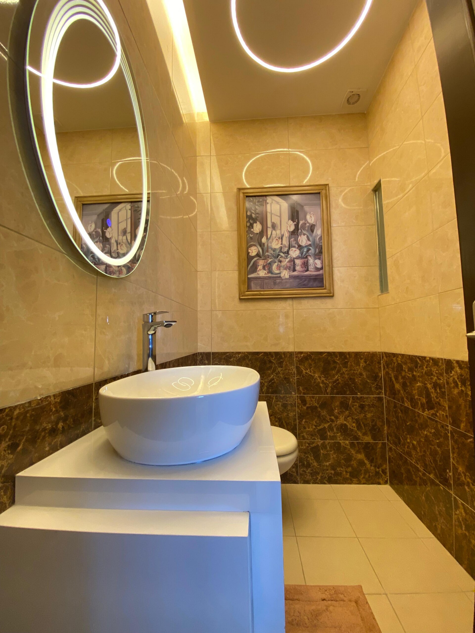 4ec9bd5c-64ac-4491-b2cd-f8c31e858e26 Modern bathroom vanity with a circular backlit mirror above a white vessel sink on a white countertop, beige tiled walls with a darker marble band, and a framed floral painting on the wall.