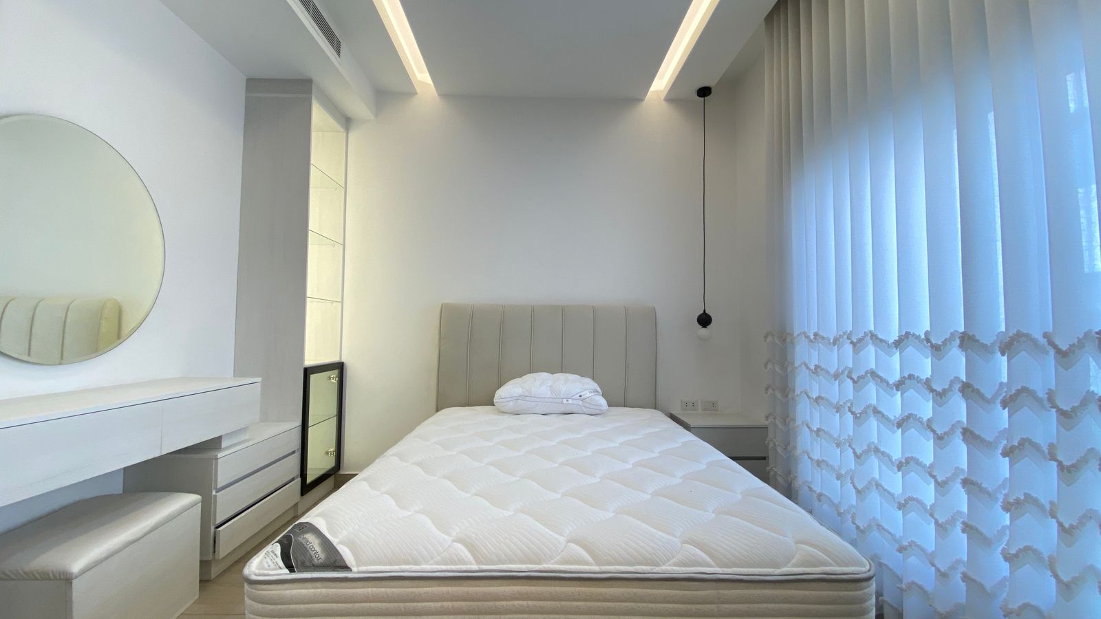 4fc3dc20-fa28-4990-a8ae-36acf5cf1849 A modern white bedroom with a padded headboard bed, folds of bedding on top, and sheer vertical blinds on the right side.