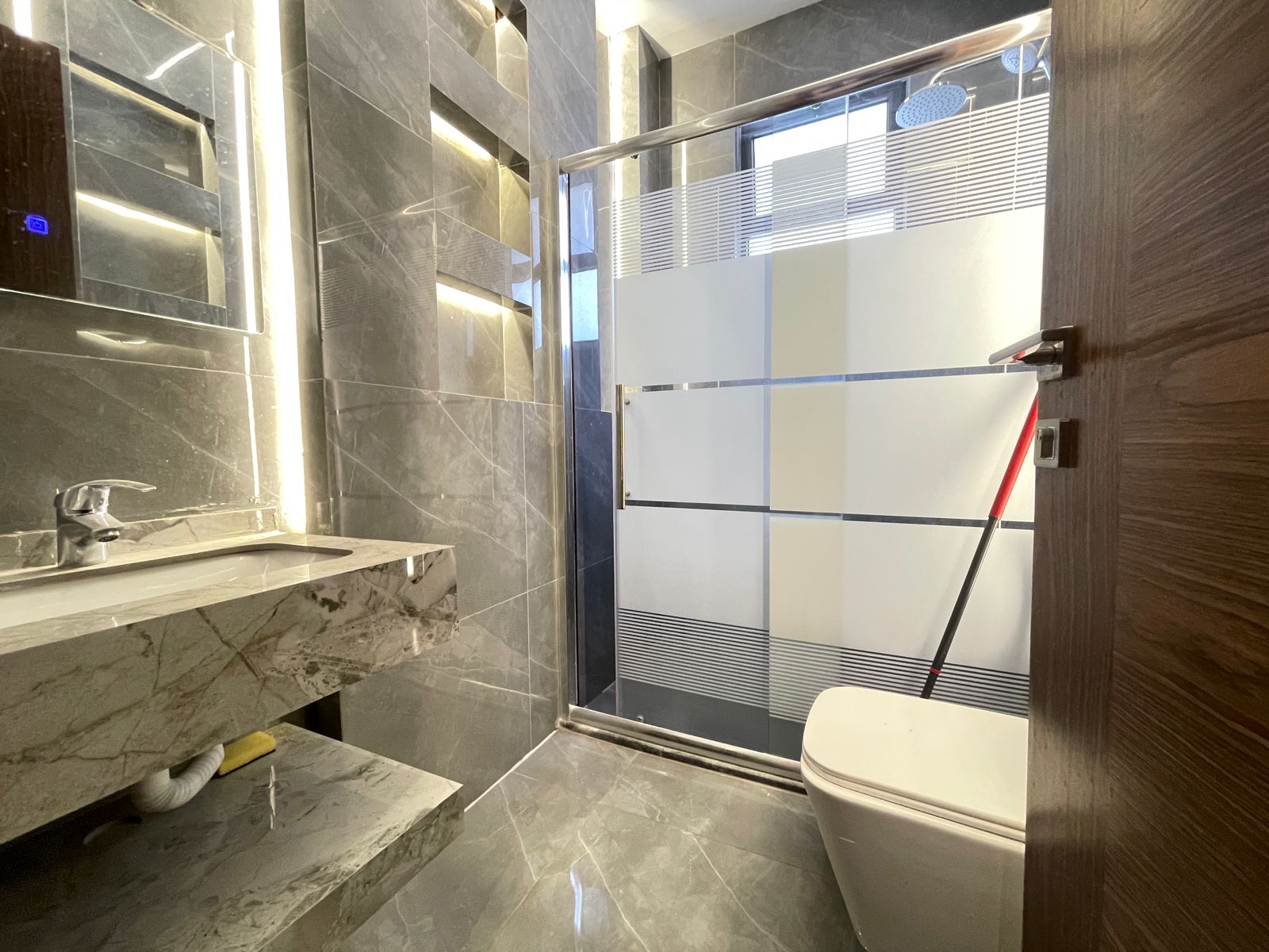 4fd73f67-aea5-47fe-a944-52201413a61d Modern grey marble bathroom with a marble vanity, backlit mirror, and glass shower enclosure; toilet on the right.