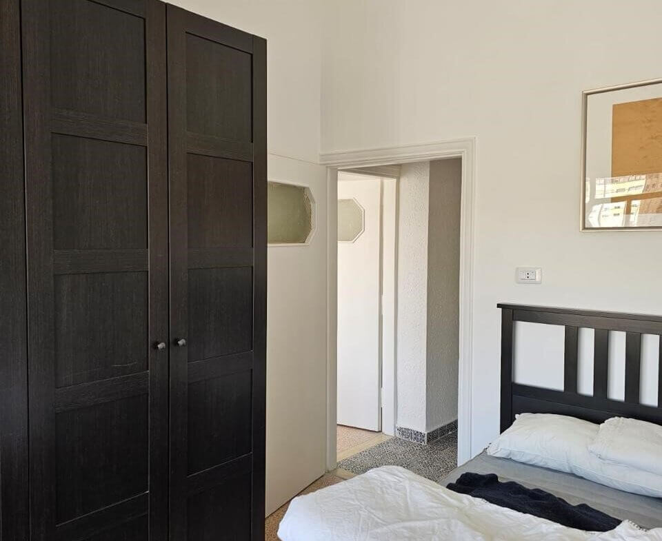 5005f9a5-e326-46ce-8f74-e9bef182454c Bedroom with a tall dark wooden wardrobe, a bed with white bedding, and an open doorway to a hallway on the right.