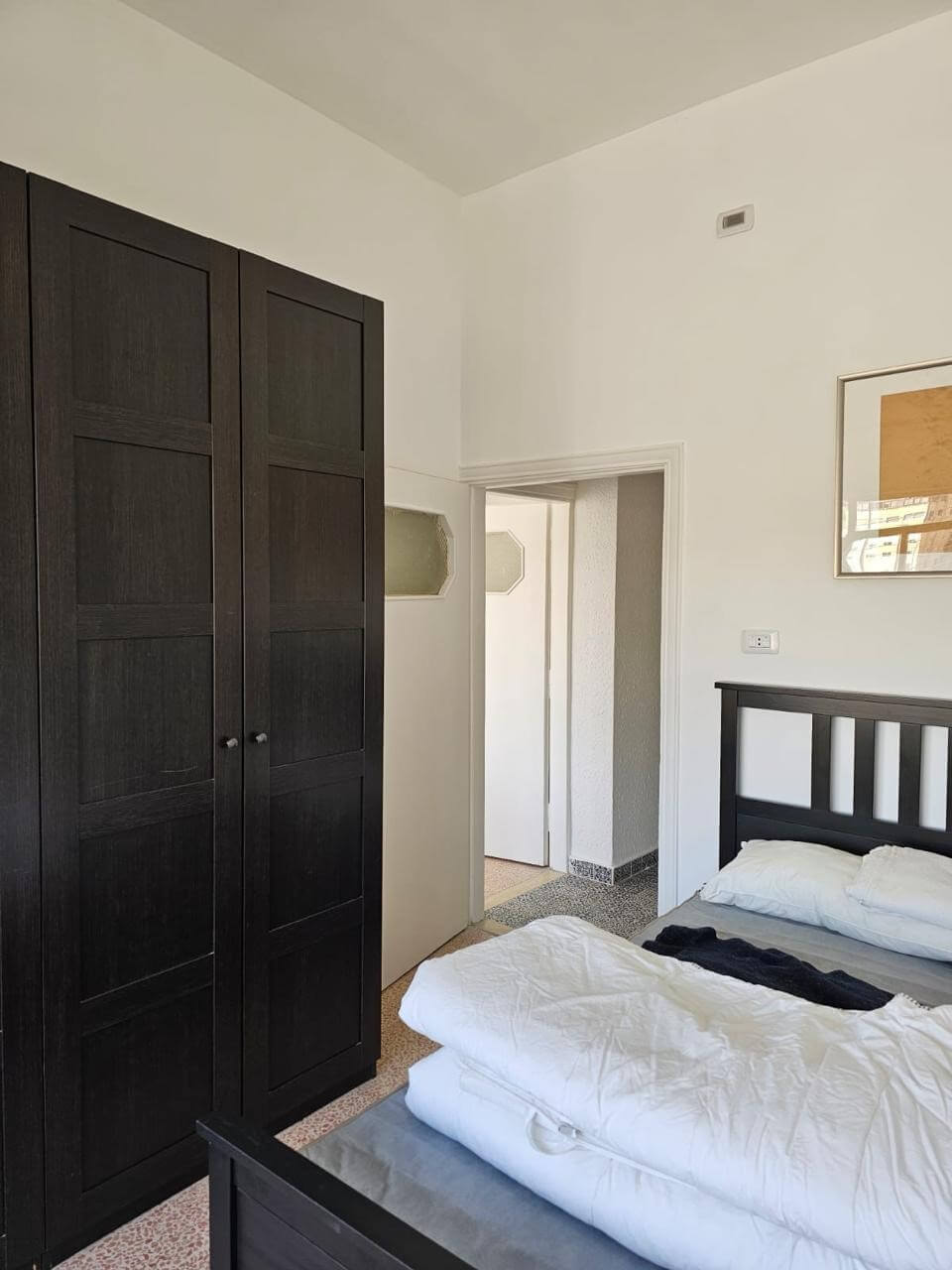 5005f9a5-e326-46ce-8f74-e9bef182454c Bedroom with a tall dark wooden wardrobe, a bed with white bedding, and an open doorway to a hallway on the right.
