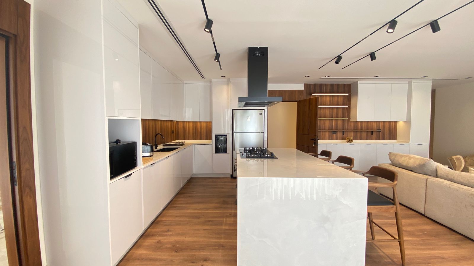 5036d1fd-420b-4d75-b0ac-affb3fea94bb Open-concept kitchen with white glossy cabinets, wood backsplash, and a large marble island with a gas cooktop and stainless fridge nearby.