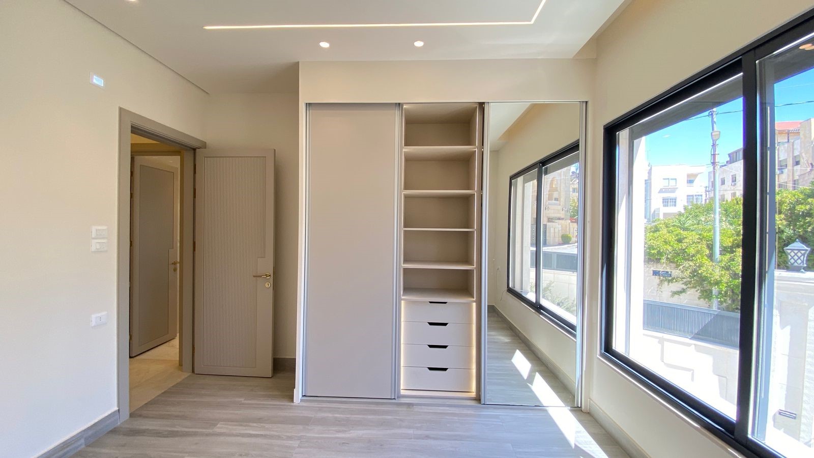 50510bcb-52a1-4d3a-a8a9-5bb826f91aa5 Modern dressing area with a built-in wardrobe (sliding doors), open shelves and four drawers, sunlit by large windows on the right, and a doorway to another room on the left.