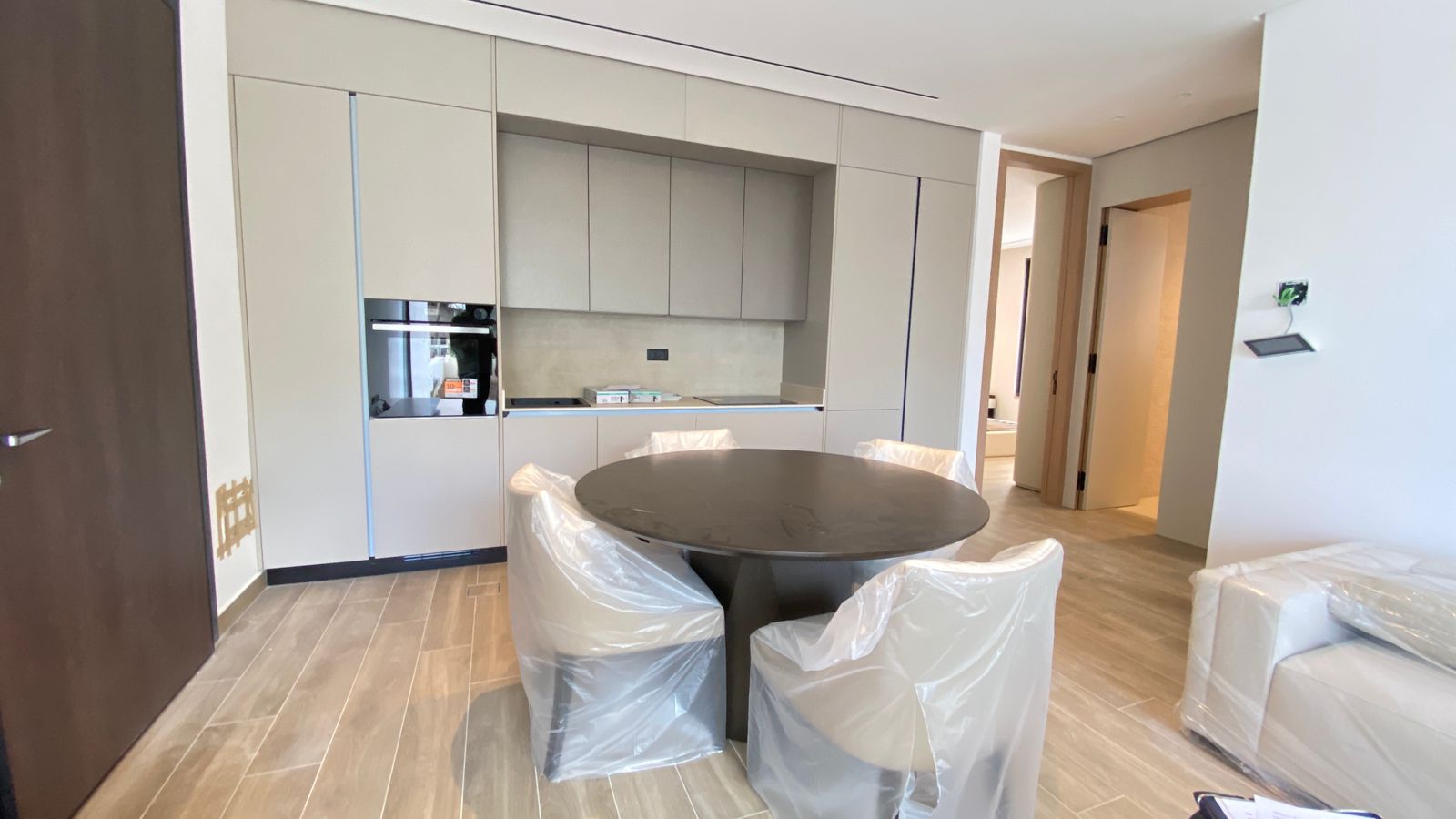 506f7ab3-ccf1-43d2-8f62-d29e1f80c269 Modern kitchen with grey cabinets, built-in oven, and a round black dining table surrounded by plastic-covered chairs.
