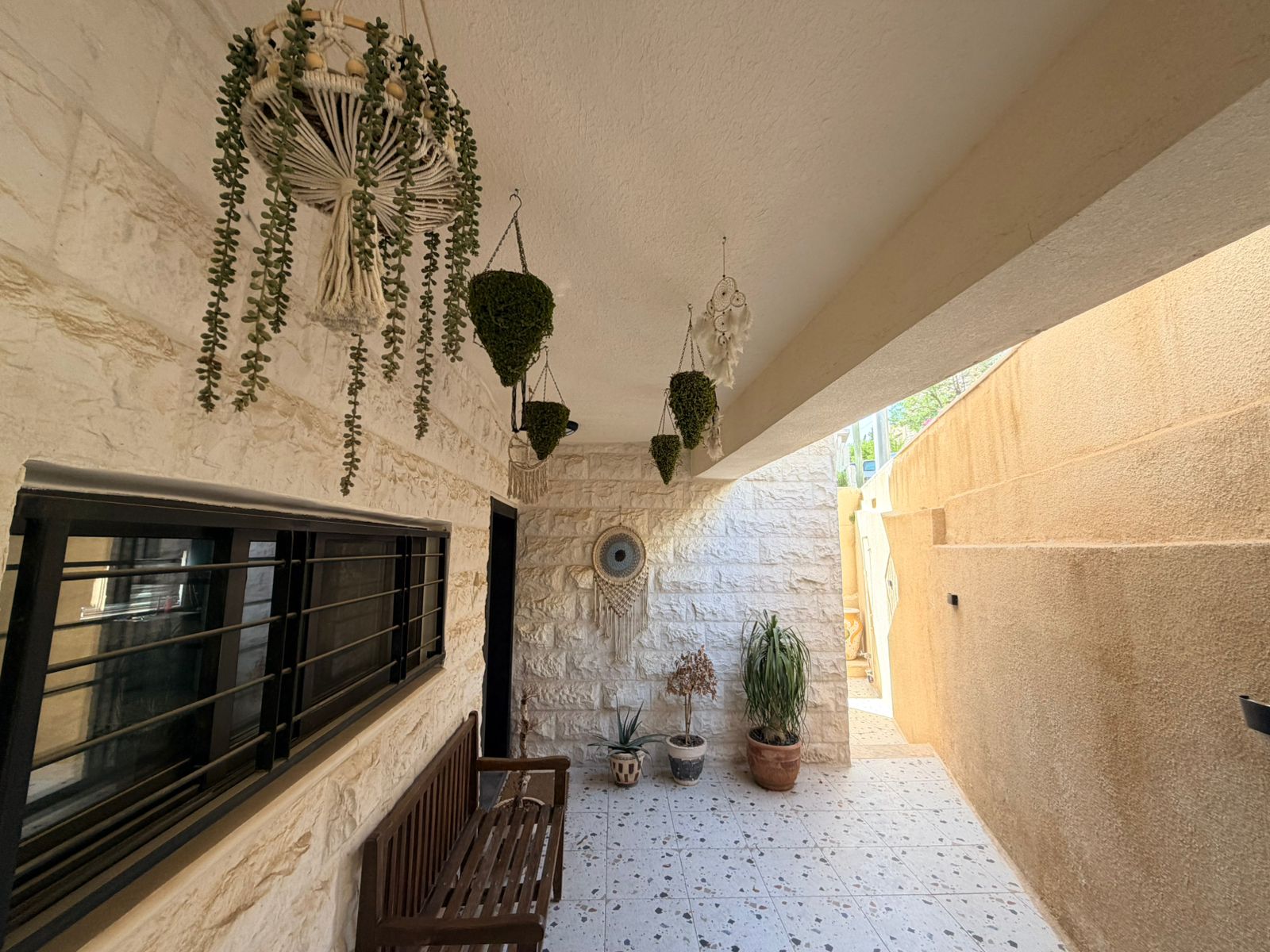 508b85c4-24c8-4c5e-a8a2-49dc7183363f Covered patio with white stone walls, row of hanging plants in macrame and several potted plants along the floor of the walkway.