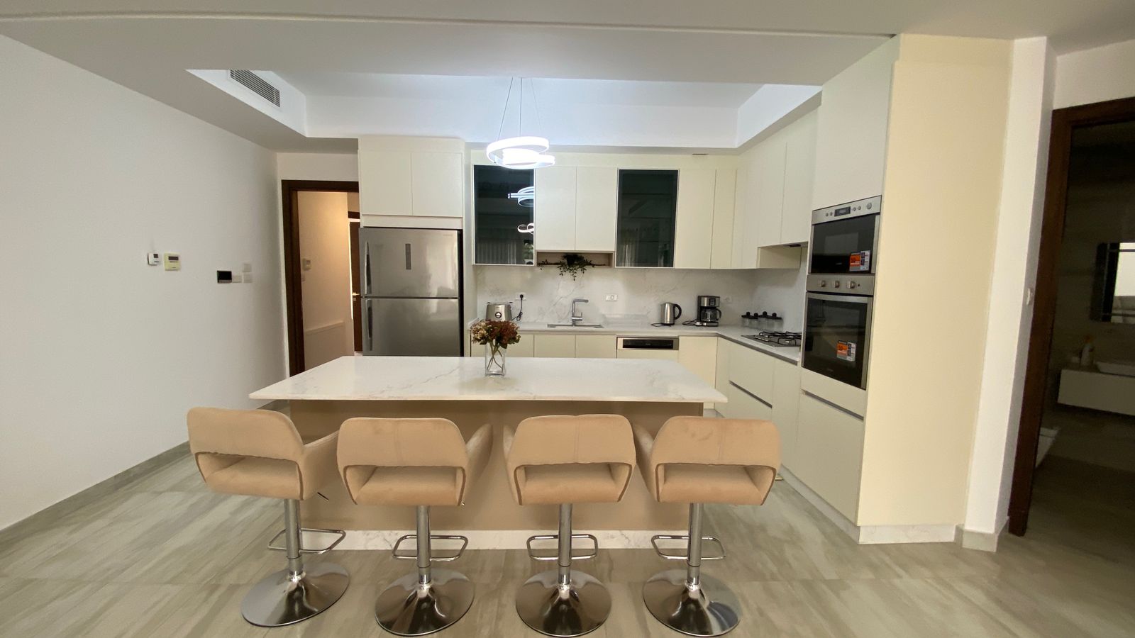 518061ab-88c9-439b-879f-ab000d6fab88 Modern kitchen with a marble island, four beige bar stools, white cabinets, stainless steel appliances, and a circular pendant light.