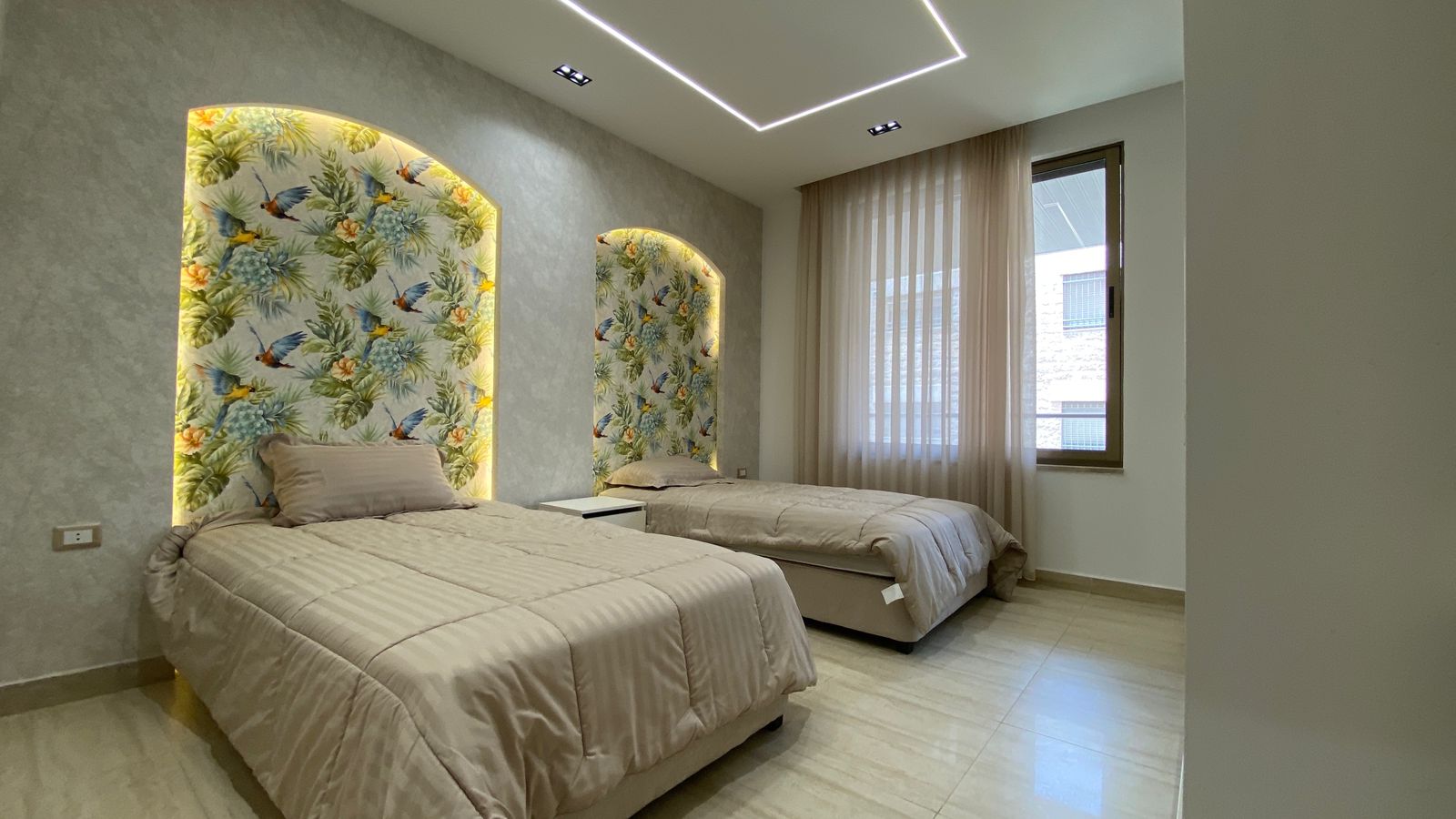 5184c32f-aa38-47f0-8e4a-54417719947e Twin beds with beige quilts in a modern room featuring tropical wallpaper arches and warm backlit edges.