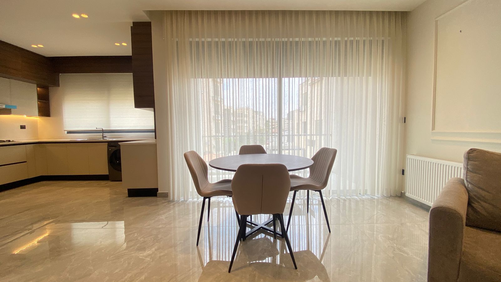524fb3ac-09d8-4b9e-9162-527f8c416684 Round dining table with four beige upholstered chairs in a bright open-plan living area, sheer curtains covering a large window beyond.