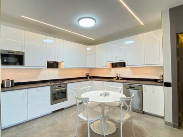 Bright modern kitchen with white glossy cabinets, black countertops, stainless steel appliances, and a round white table with four chairs in the center. A warm ceiling light and linear LED strips illuminate the space.