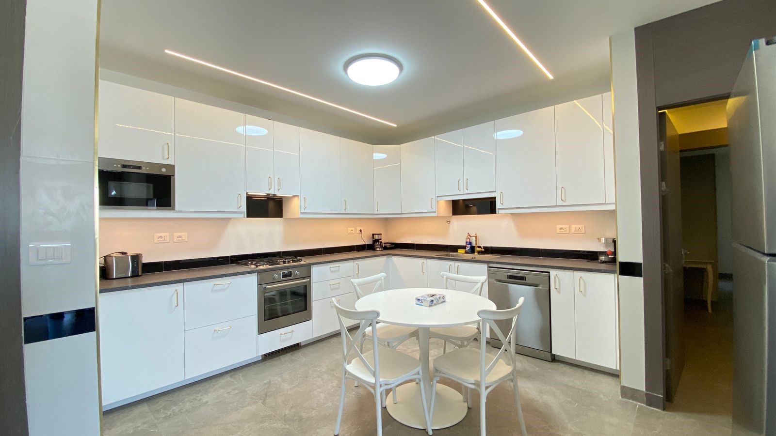 52fa8067-869d-4cbf-b517-87ebb2f22361 Bright modern kitchen with white glossy cabinets, black countertops, stainless steel appliances, and a round white table with four chairs in the center. A warm ceiling light and linear LED strips illuminate the space.