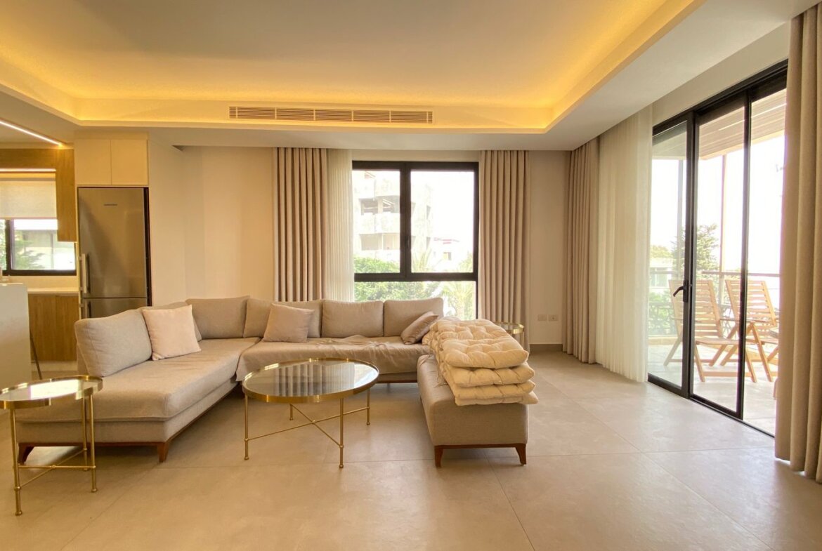 Open-plan living room with a curved L-shaped beige sofa, round glass coffee tables, and beige curtains.