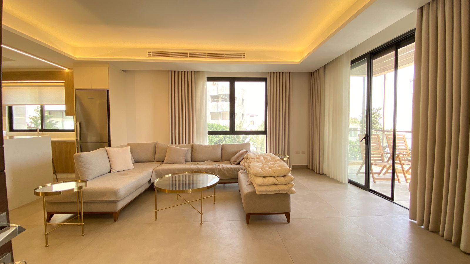 53ced26c-8fb7-4883-8bd8-85f53947d3e8 Open-plan living room with a curved L-shaped beige sofa, round glass coffee tables, and beige curtains.