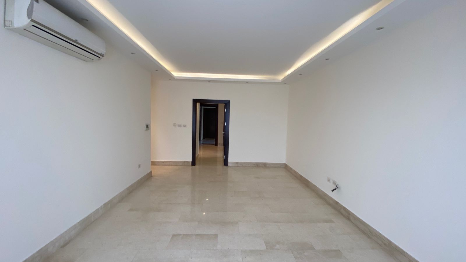53d51182-d65d-4ded-a921-2941e760c323 Empty modern living room with white walls, beige marble floor, recessed ceiling lights, and a wall-mounted air conditioner on the left; doorway visible at the far end.