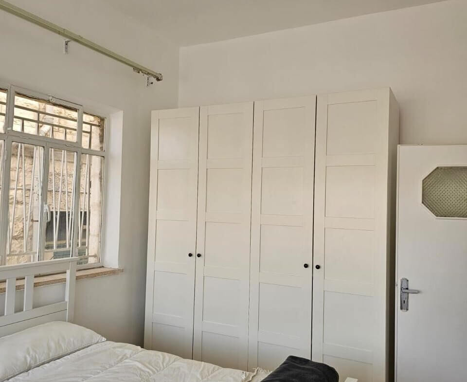 5426d401-d876-42e0-9546-8262bf4b562c White bedroom with a tall white wardrobe, a white bed, and a dark folded blanket at the foot, window to the left with bars.