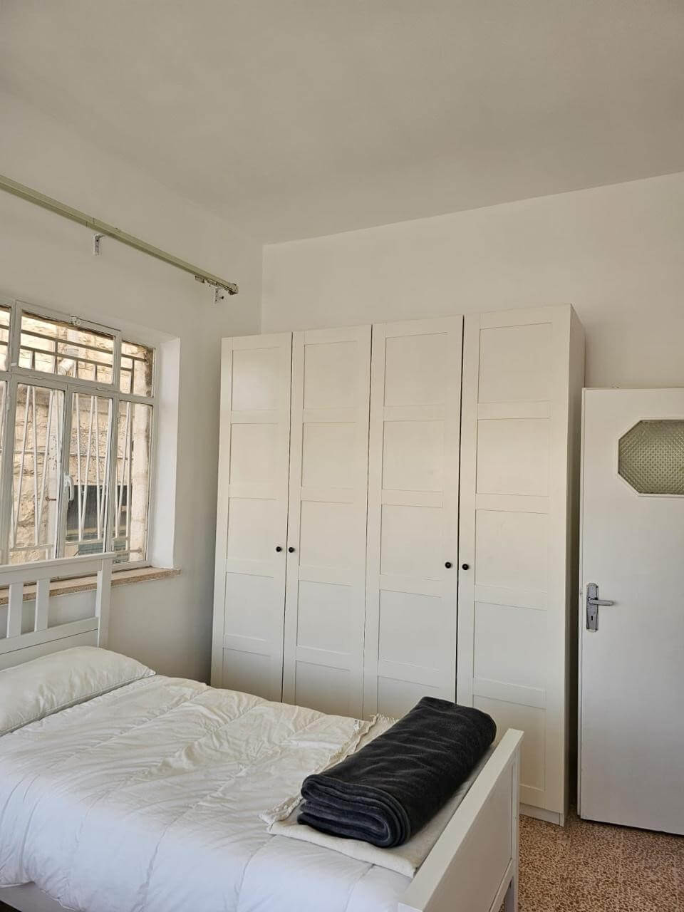 5426d401-d876-42e0-9546-8262bf4b562c White bedroom with a tall white wardrobe, a white bed, and a dark folded blanket at the foot, window to the left with bars.